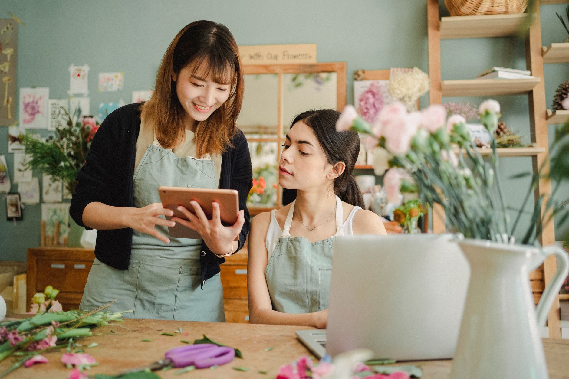 Two coworkers in aprons at a table with fresh flowers, looking at a tablet together