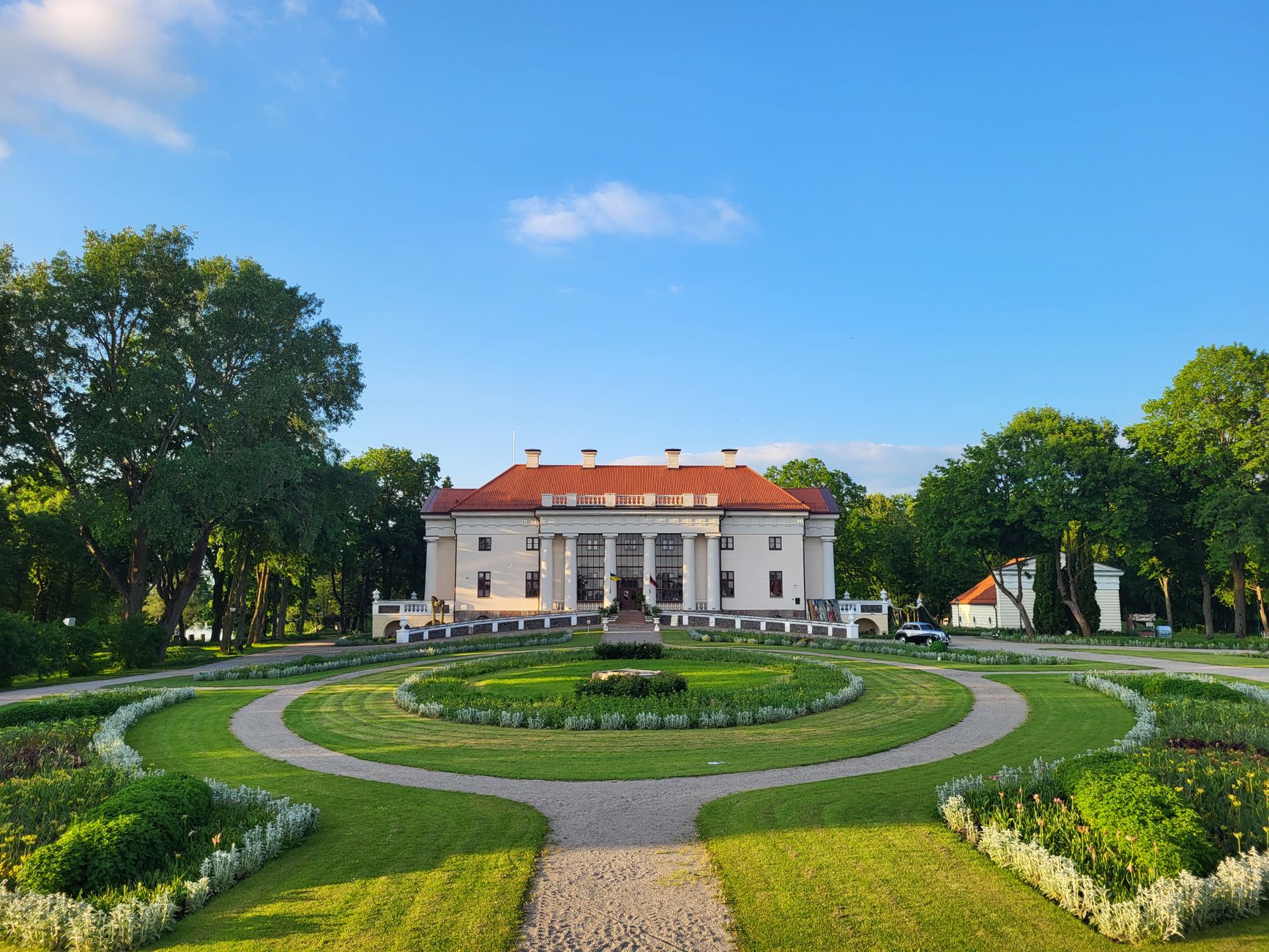 A white mansion beyond a symmetrical estate garden of trimmed lawns and paths under a blue sky