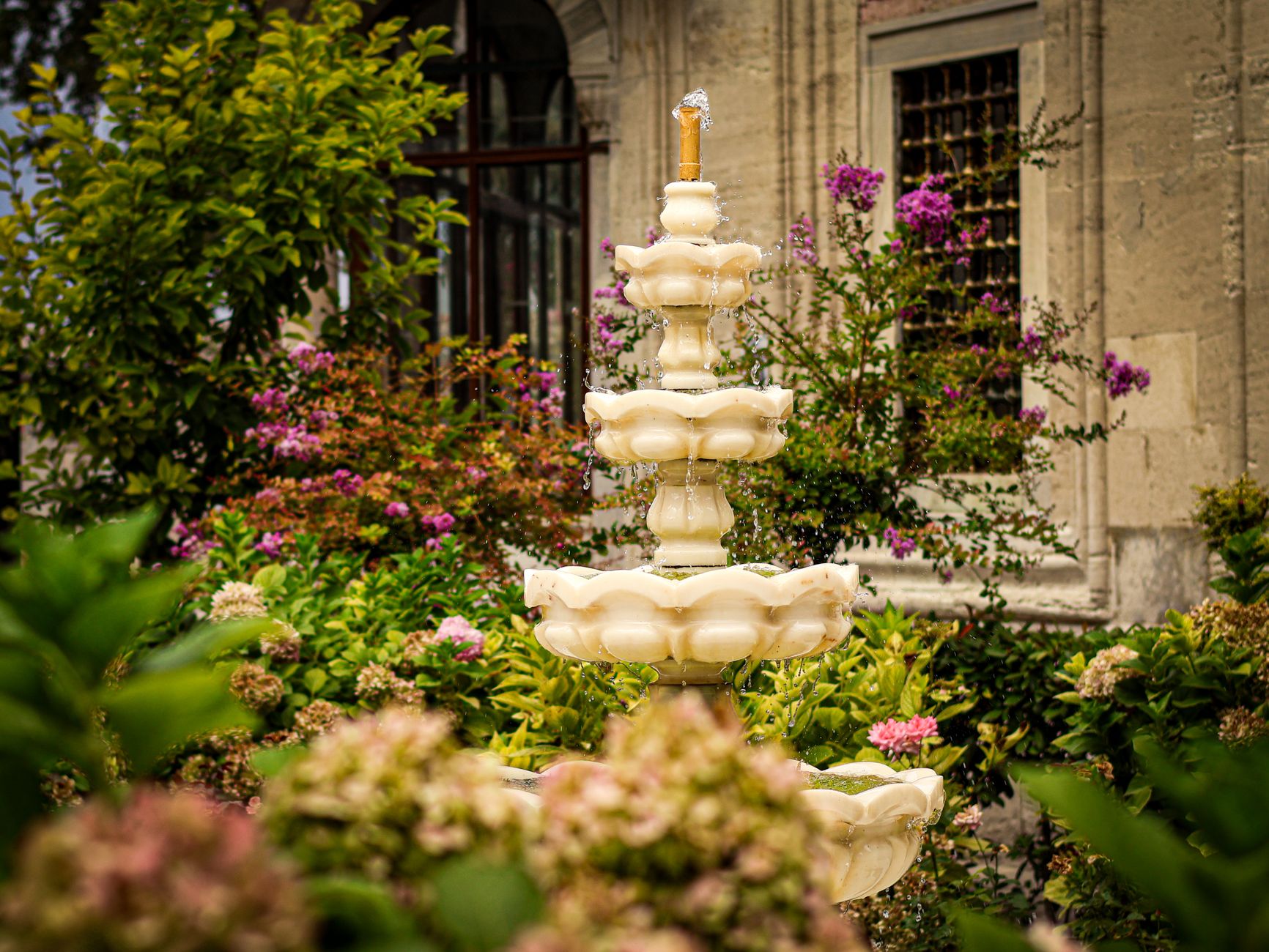 A tiered stone fountain surrounded by pink flowers and greenery in a courtyard garden