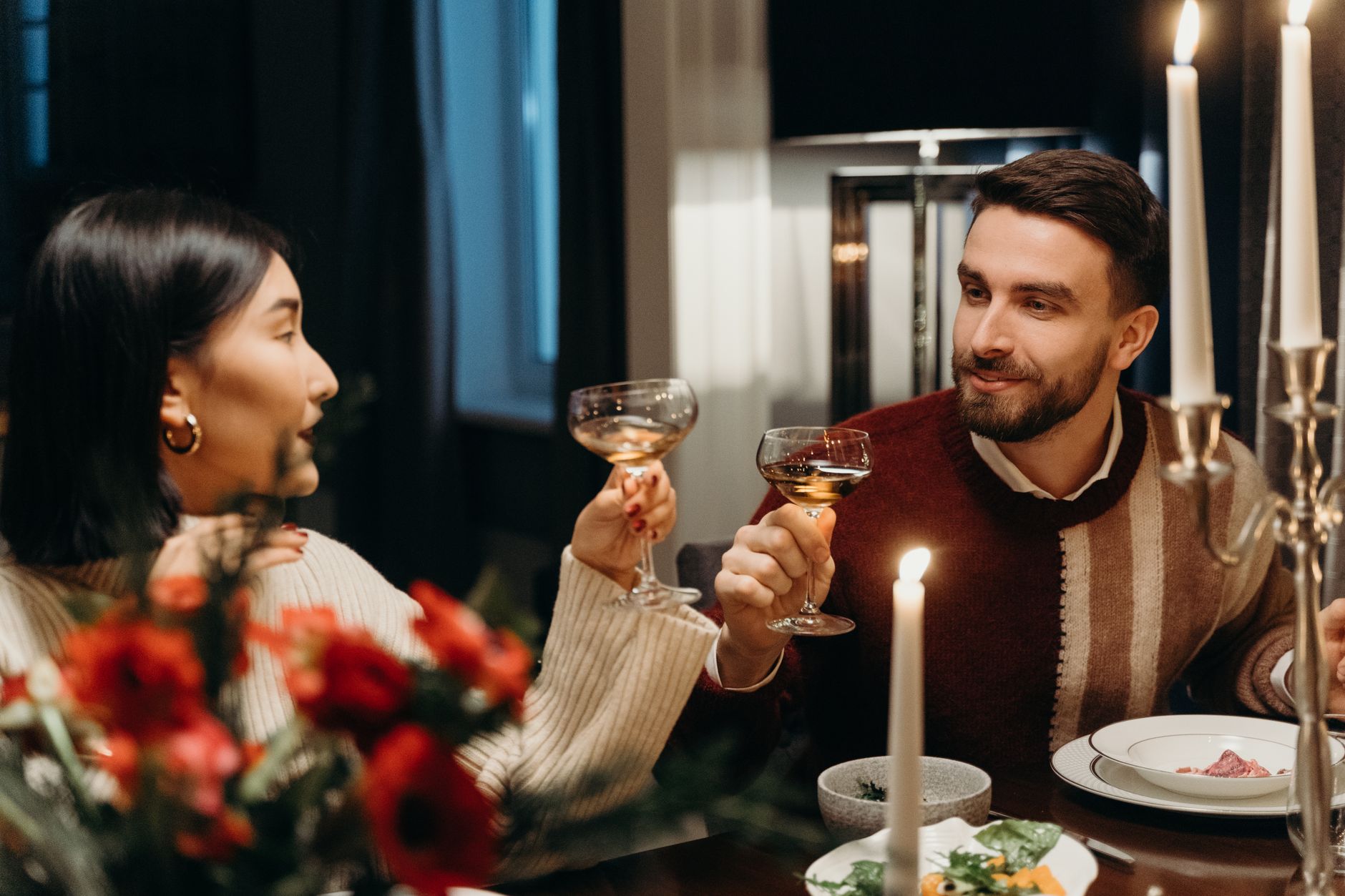 A couple toasting at a candlelit dinner table with red flowers in the foreground