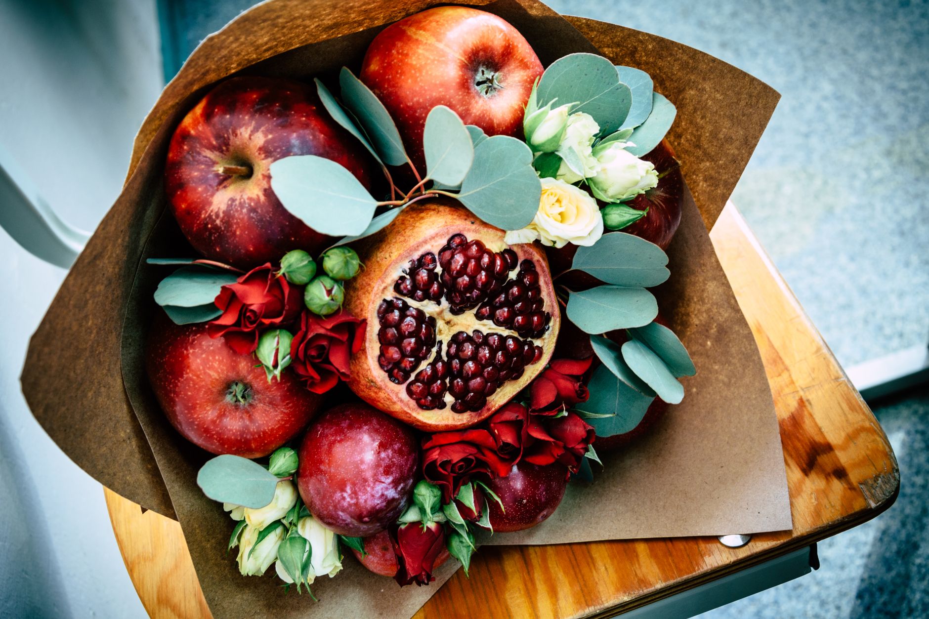 A wrapped autumn-toned arrangement of apples, a halved pomegranate, greenery, and red flowers