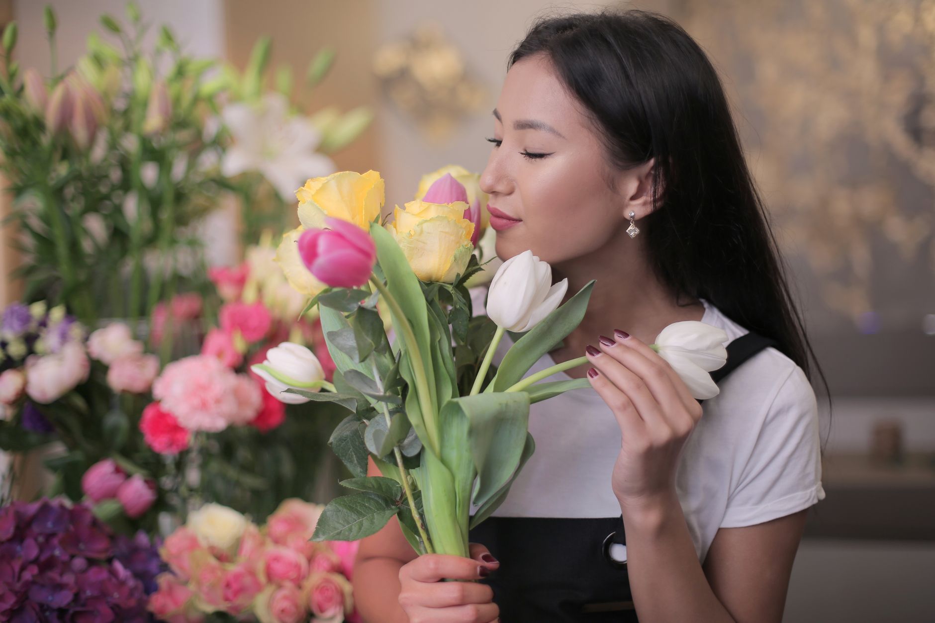 A woman holding a mixed bouquet and smelling a white tulip in a flower-filled room