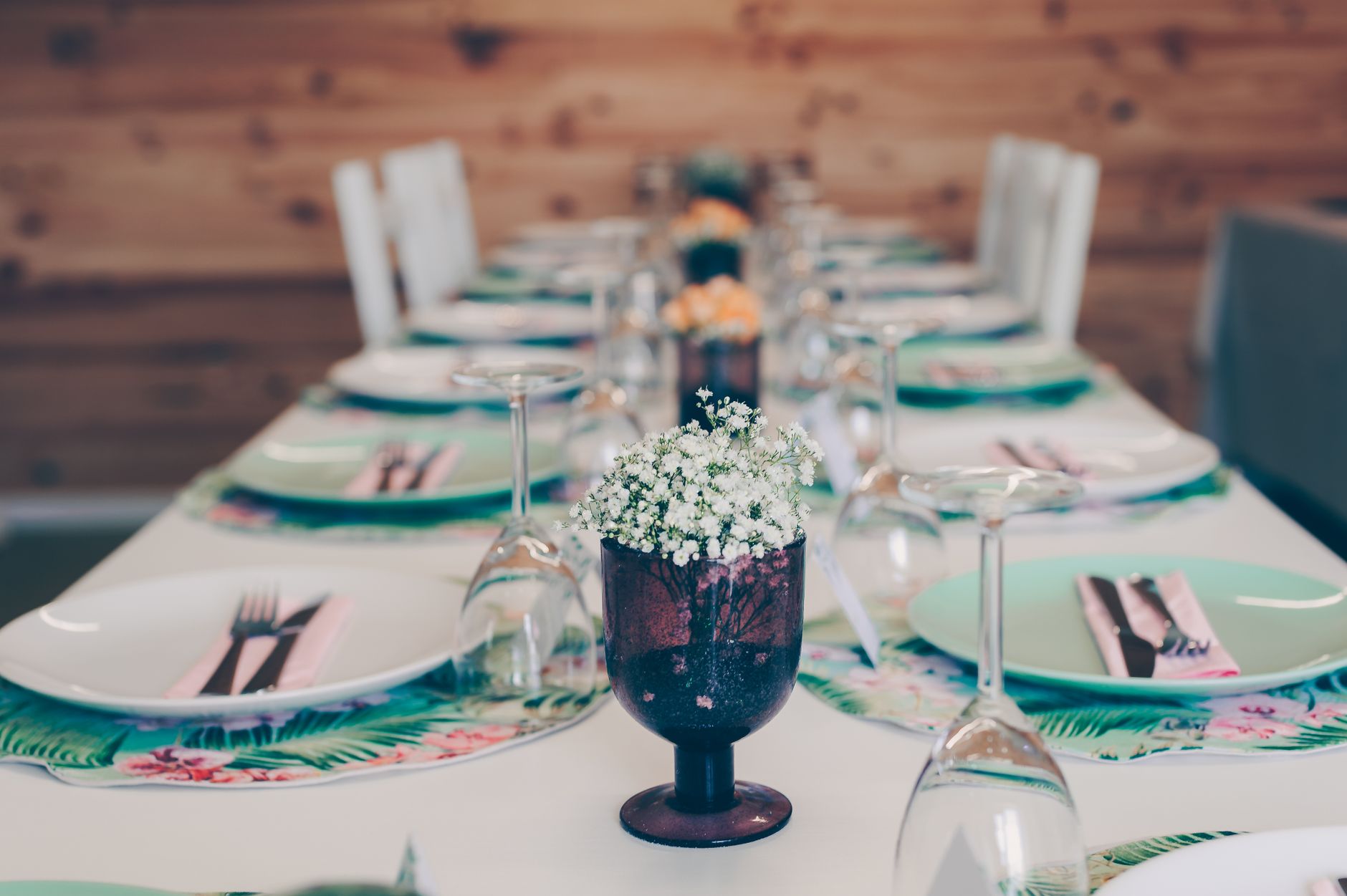 A long dining table set with plates and wine glasses with a small white floral centerpiece in front