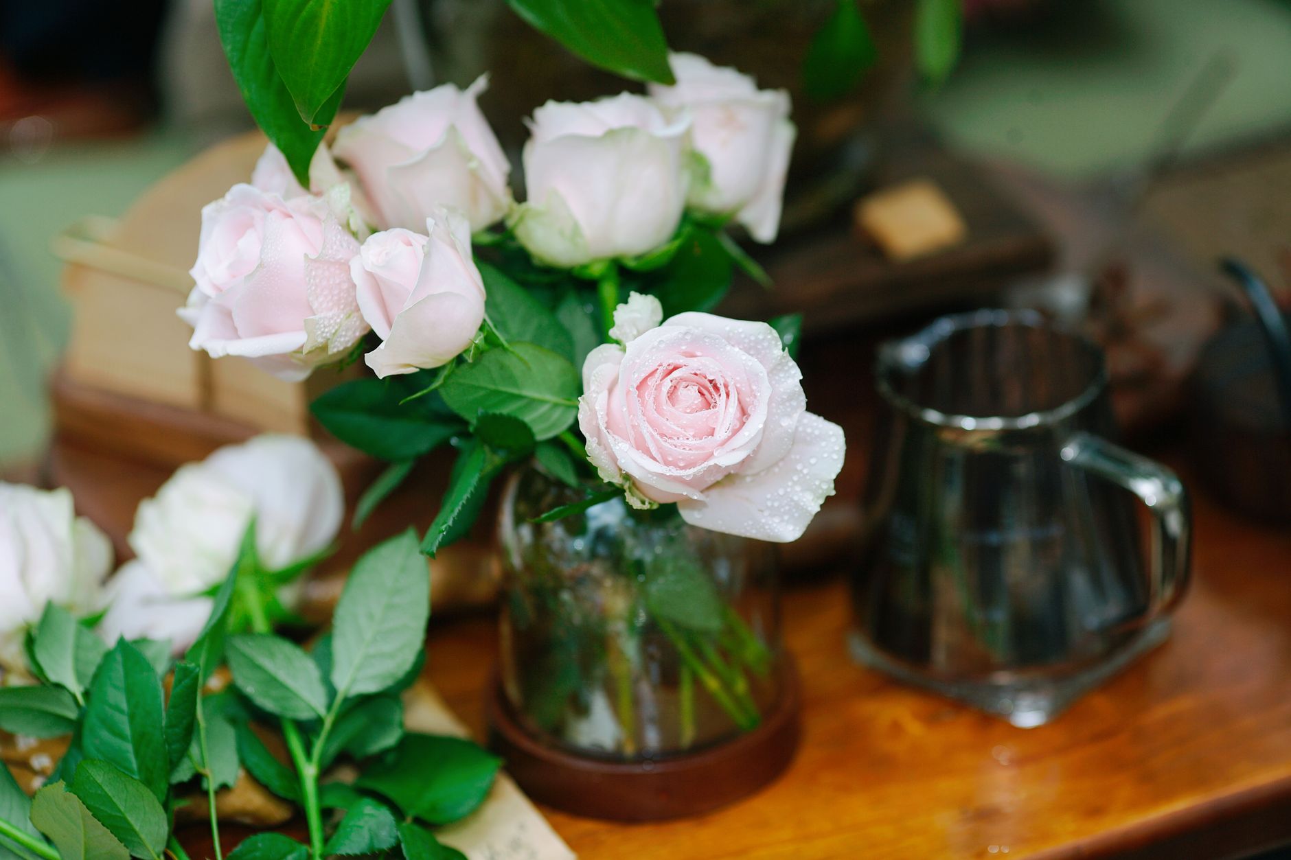 Pale pink roses in a glass vase on a rustic wooden table beside a metal pitcher