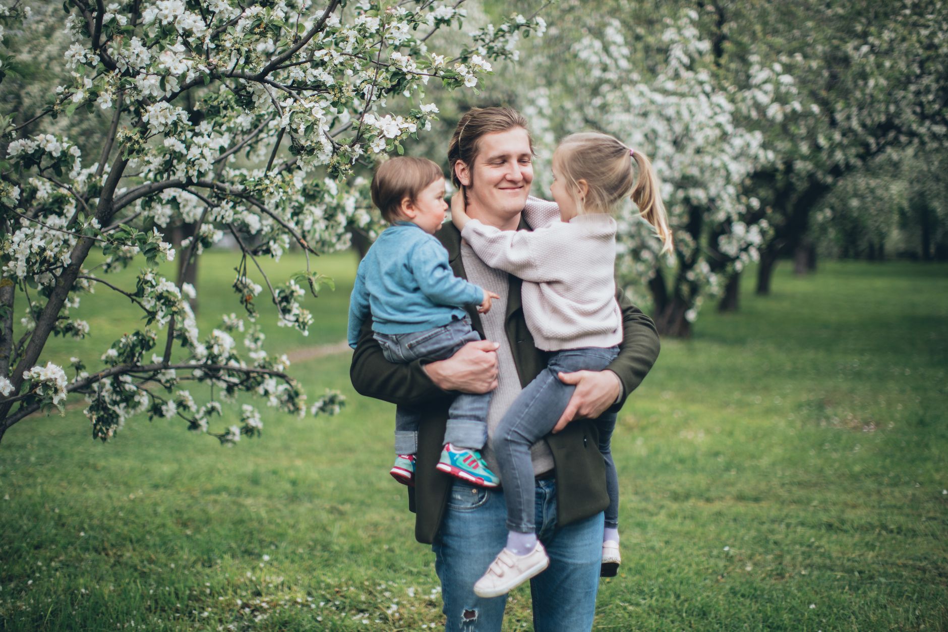 A father holding his two children beneath blooming spring trees in a grassy park
