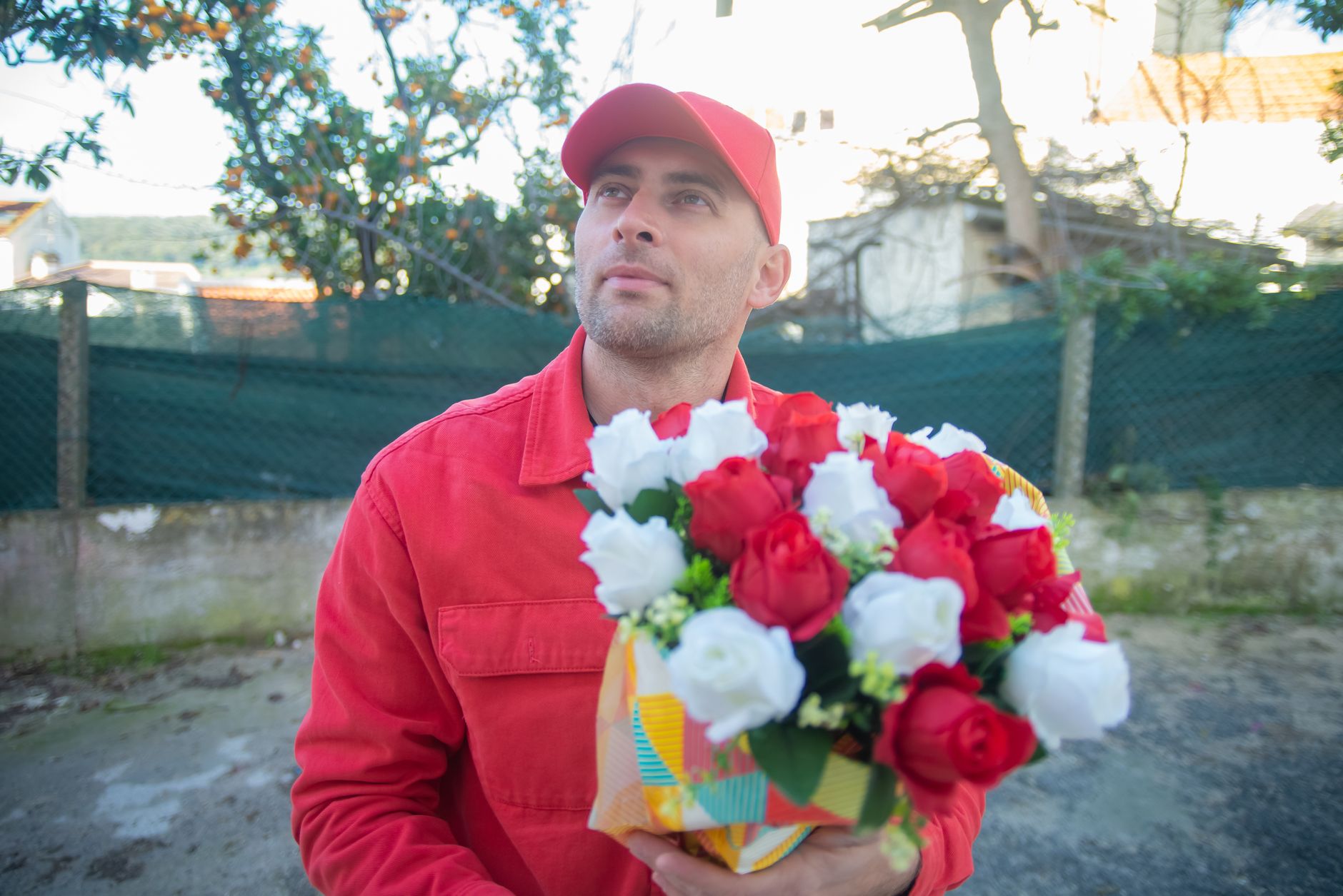 A delivery man in a red cap carrying a wrapped bouquet of red and white flowers outdoors