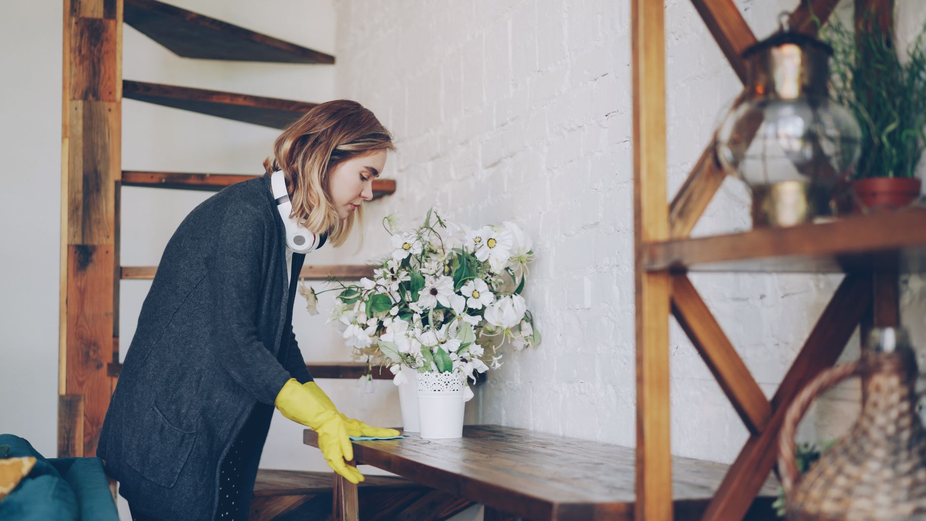 A woman wearing yellow gloves wiping a wooden table beside a white flower arrangement in a bright room