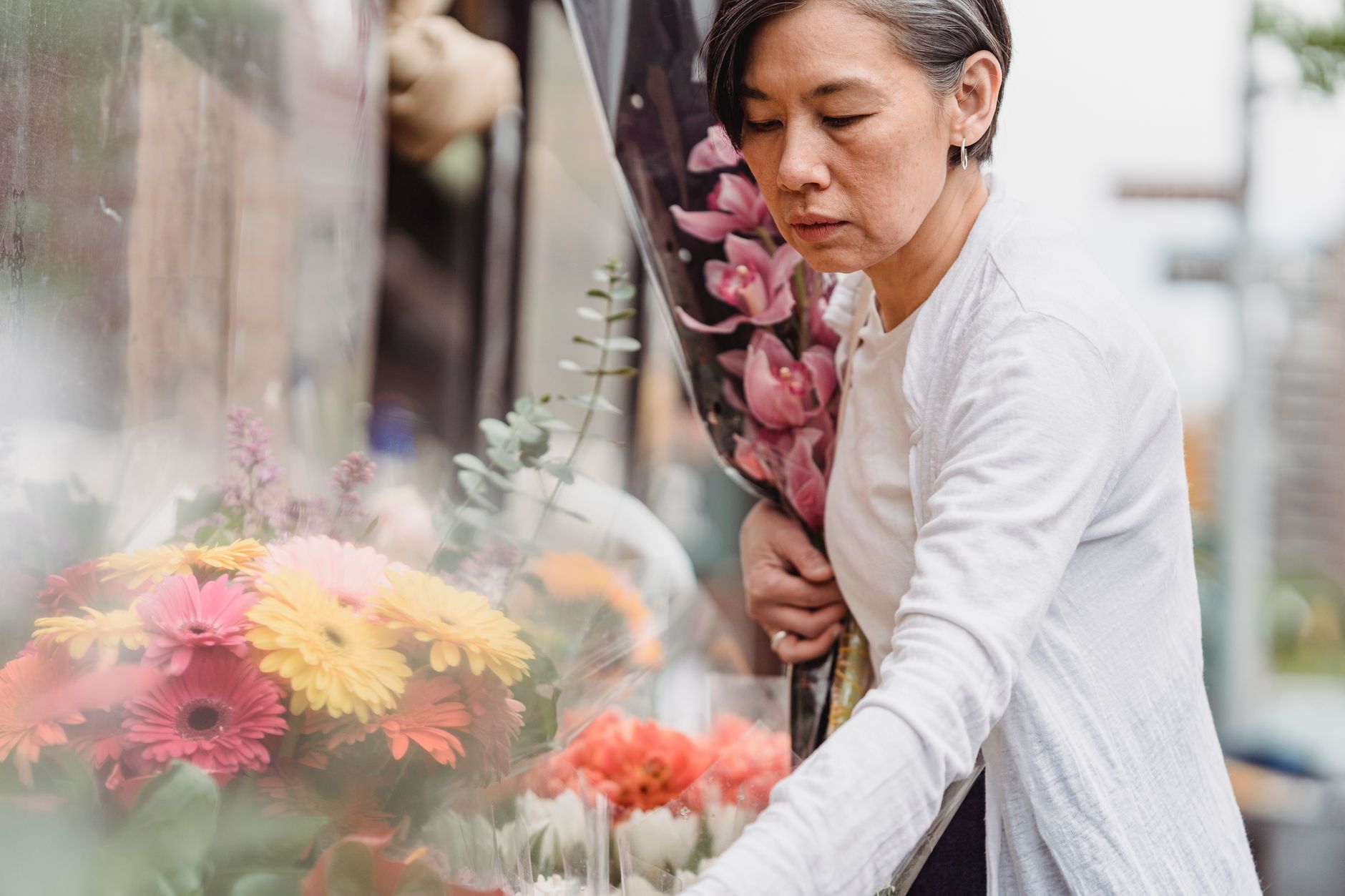 A woman in a white cardigan carefully choosing a bouquet from a flower shop display of gerberas and orchids