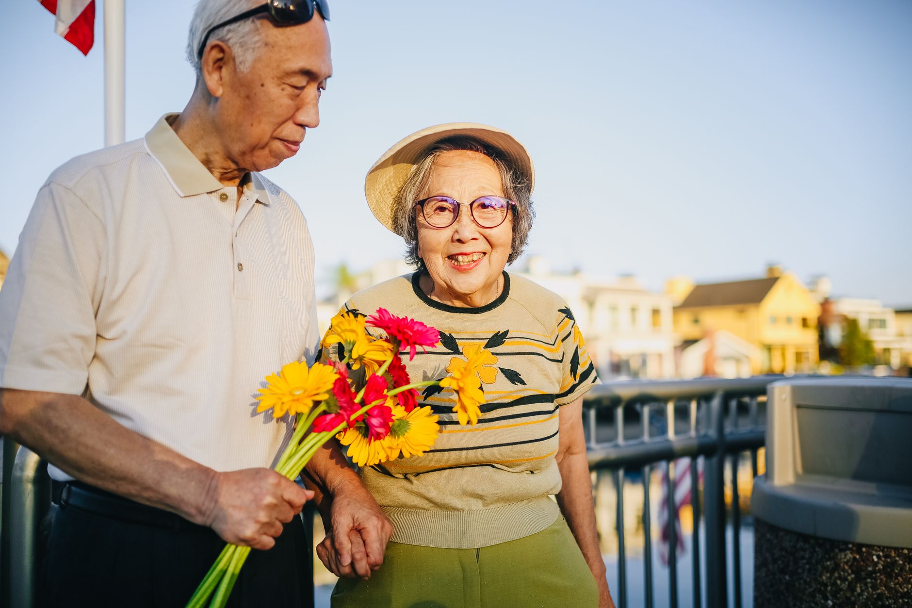 An elderly couple smiling outdoors while holding a bouquet of yellow and pink gerbera daisies