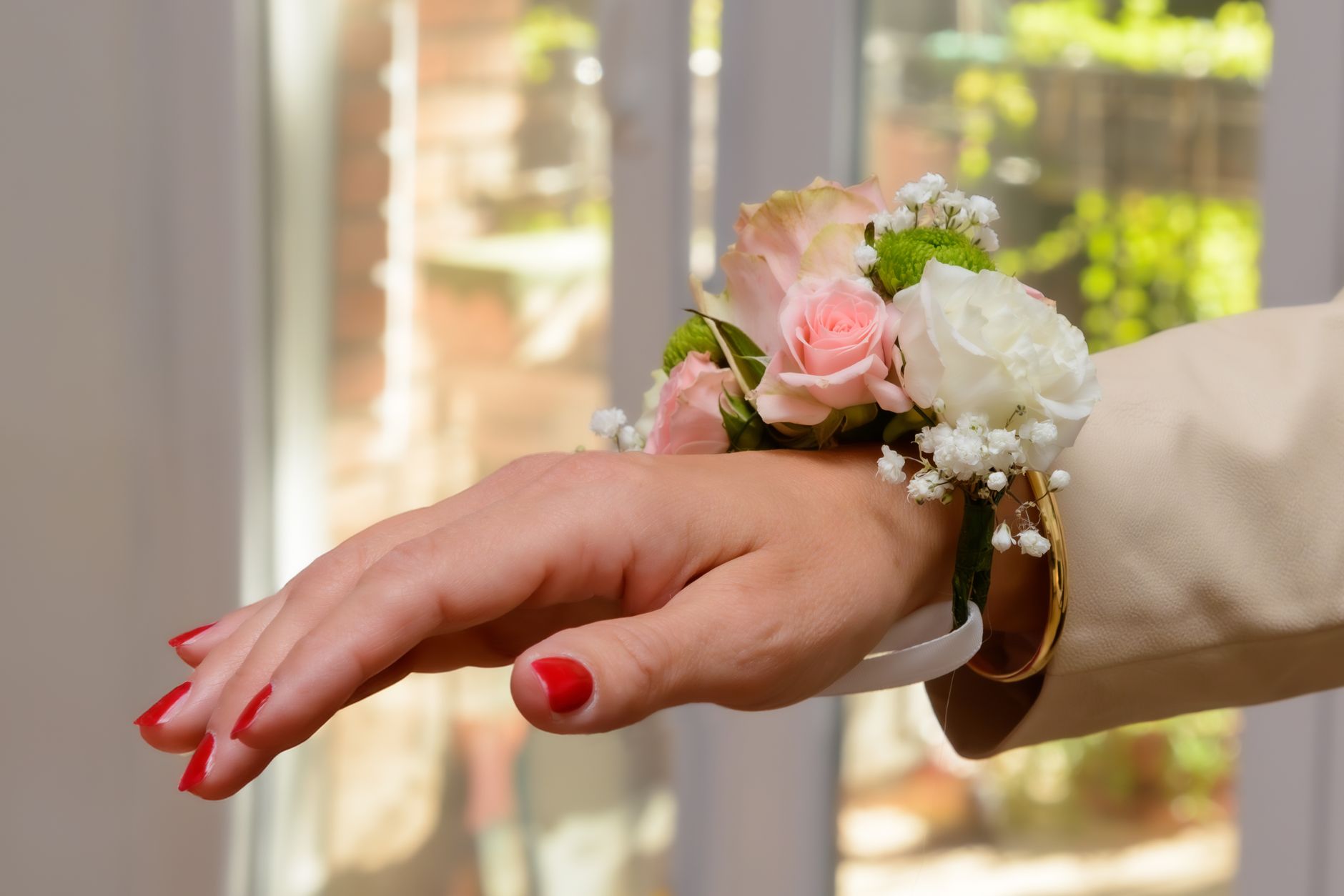 A close-up of a pink and white rose wrist corsage on a hand reaching toward a sunlit window