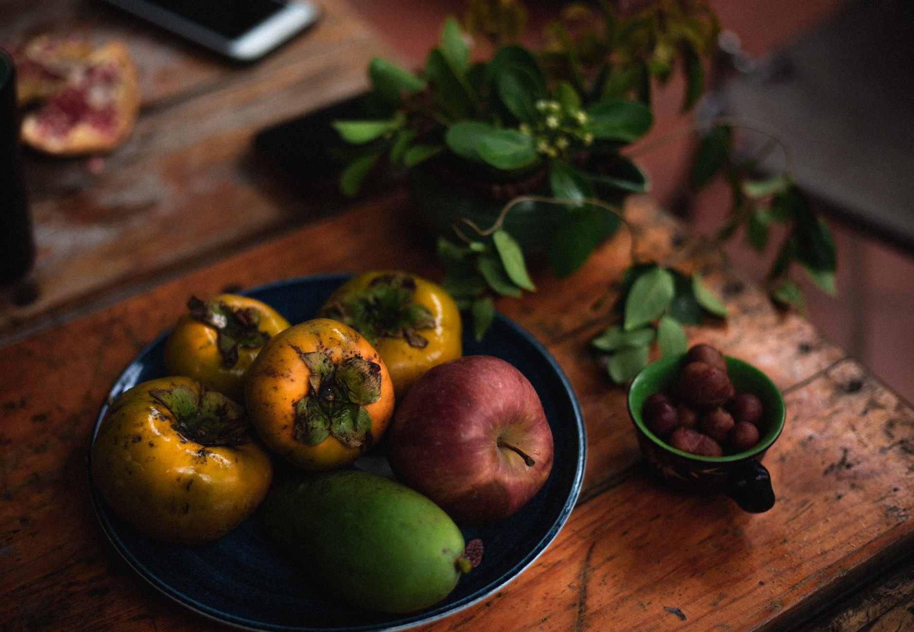 A blue plate of ripe persimmons, an apple, and a pear on a rustic wooden table beside a sprig of greenery