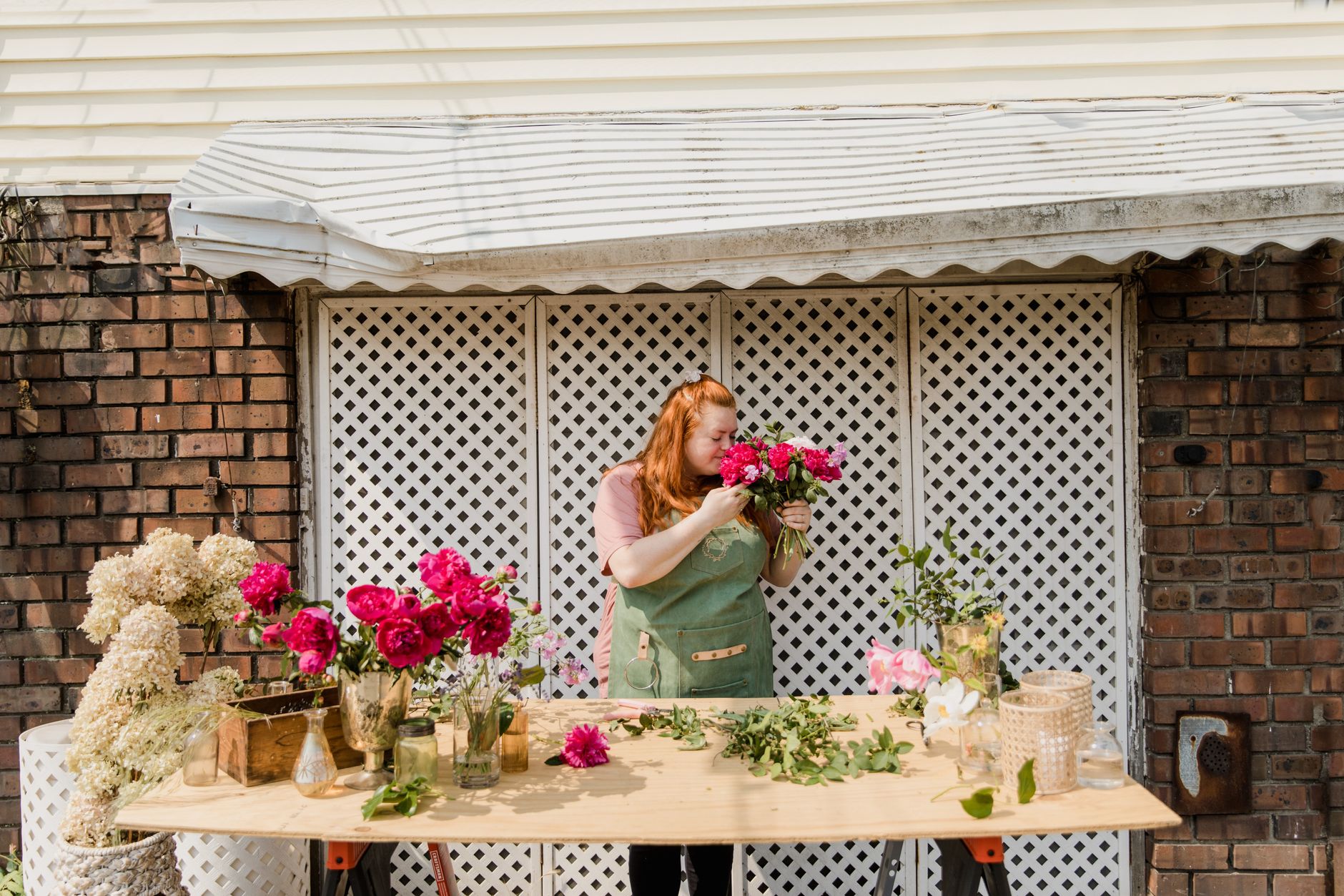 A florist in a green apron holding a bunch of pink peonies at a long wooden table of flowers outside a brick building